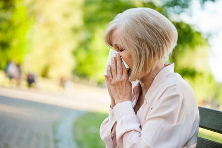 Adult woman is sitting in park and blowing nose. She is having allergy.の写真素材