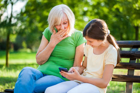 Grandmother and granddaughter are sitting in park and using mobile phone.の写真素材