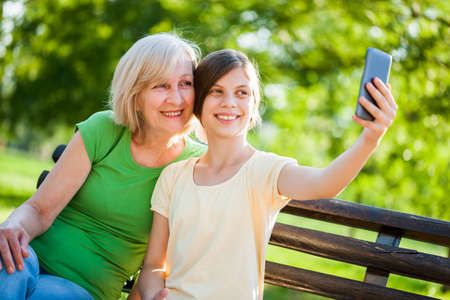 Grandmother and granddaughter are sitting in park and taking selfie.の写真素材