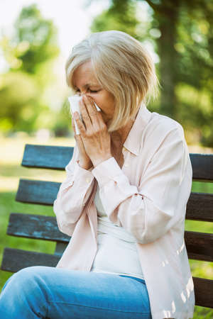 Adult woman is sitting in park and blowing nose. She is having allergy.の写真素材