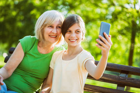 Grandmother and granddaughter are sitting in park and taking selfie.の写真素材