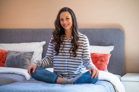 Happy young woman sitting on bed in her home, looking at camera and smiling.の写真素材