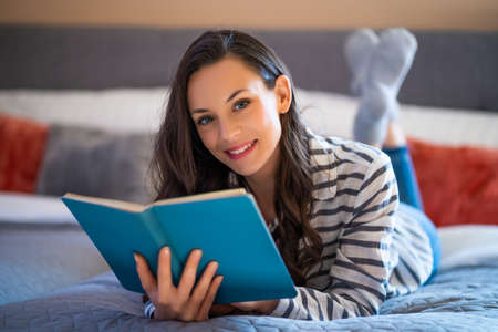 Happy young woman lying on bed in her home and reading book.の写真素材