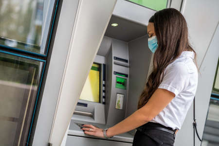 Businesswoman with protective mask is using ATM machine to withdraw cash. Covid-19 virus pandemic concept.の写真素材