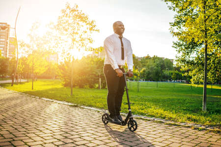 Happy african-american businessman is riding scooter in park on sunny day.の写真素材
