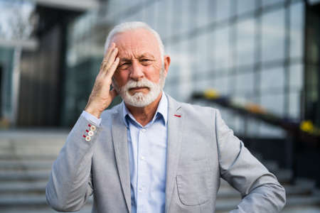 Senior businessman is tired of work. He is standing in front of company building and having headache.の写真素材
