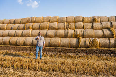 Happy farmer is standing beside bales of hay. He is satisfied because of successful harvesting.の写真素材