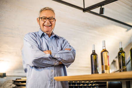 Portrait of happy senior man who owns winery. He is standing in his wine cellar. Industry wine making concept.の写真素材