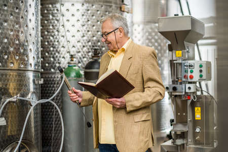 Portrait of happy senior man who owns winery. He is standing beside wine storage tanks and examining...