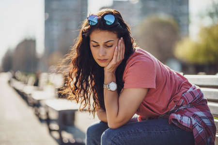 Sad young woman is sitting on bench in city. She is depressed.
