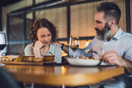 Adult couple is sitting at restaurant. They are having dinner with wine. Woman is sad and depressed.