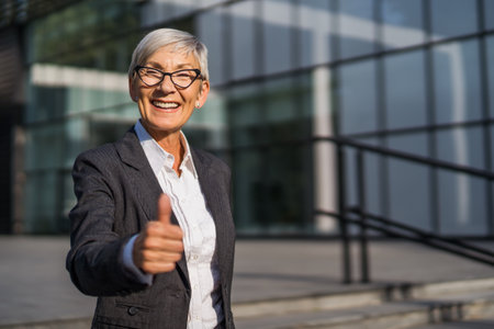 Outdoor portrait of successful senior businesswoman in front of company building. She is looking at camera and showing thumbs up.の写真素材