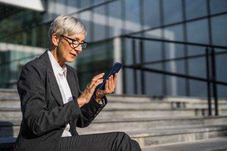 Senior businesswoman sitting in front of company building and messaging on smartphone.の写真素材