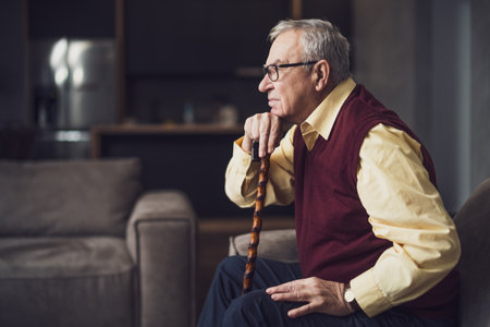 Pensive and worried senior man is sitting on bed in his home. Portrait of senior man with walking cane.の写真素材