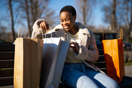 Outdoor portrait of happy black woman. She is sitting on bench in the street and looking at shopping bag.の写真素材