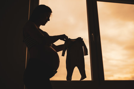 Pregnant woman standing beside window and holding baby clothes. Silhouette of pregnant woman.の写真素材
