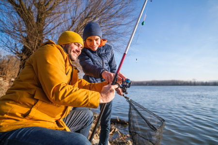 Father and son are fishing on sunny winter day. Freshwater fishing. Teenage boy is learning to fish.の写真素材