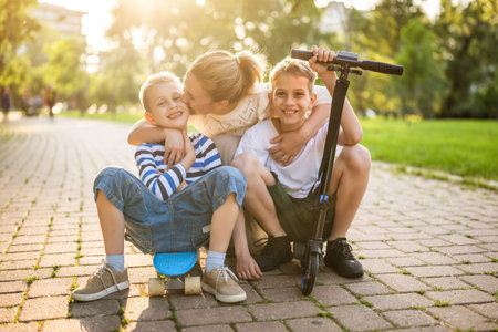 Joyful mother having fun with her two sons in park on sunny day. They are ridding skateboard and scooter.の写真素材