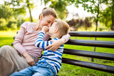 Happy mother is sitting with her son on bench in park. They are having fun together.の写真素材