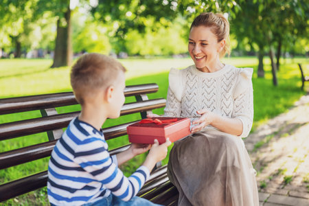 Happy mother is sitting with her son on bench in park. Boy is giving a present to his mother.の写真素材