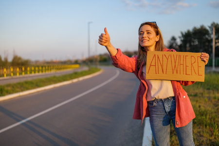 Woman is hitchhiking on roadside trying to stop car. She is holding cardboard with inscription.の写真素材