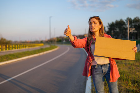 Woman is hitchhiking on roadside trying to stop car. She is holding blank cardboard for your text.の写真素材