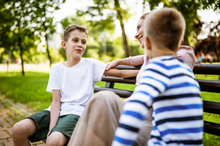 Happy mother is sitting with her sons on bench in park. They are talking and enjoying their time together.の写真素材