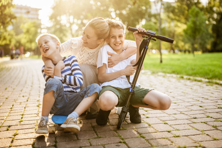 Joyful mother having fun with her two sons in park on sunny day. They are ridding skateboard and scooter.の写真素材