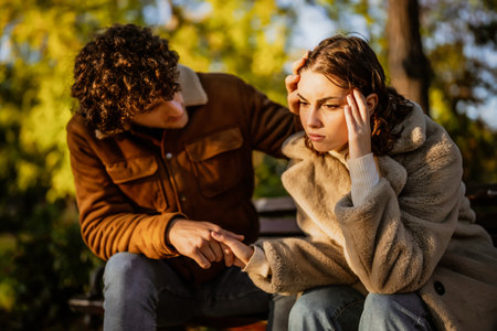Young couple is sitting in park on sunny day. Woman is sad and man in consoling her.の写真素材
