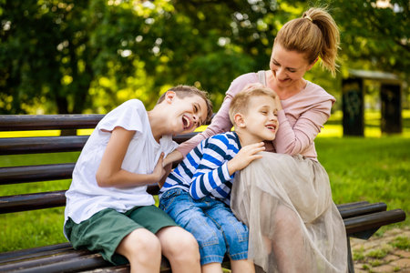 Happy mother is sitting with her sons on bench in park. They are having fun together.の写真素材