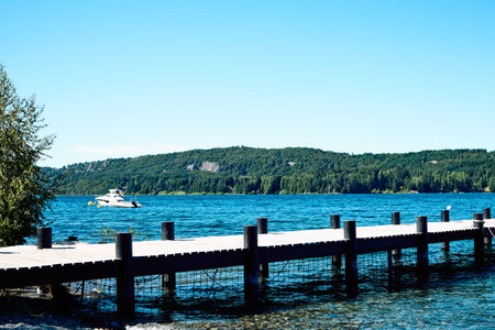 Beautiful mountain lake. Transparent blue water. Shore pier. Surrounded by rocky mountains and forest. In the distance there is a yacht on the waves. Bright green trees and shrubs bordering a mountain lake.の写真素材