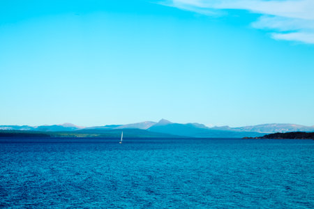 Beautiful mountain lake. Transparent blue water. Surrounded by rocky mountains and forest. In the distance a sailboat on the waves.の写真素材