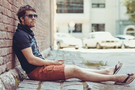 Bearded young stylish hipster man using phone sitting next to a brick wallの写真素材