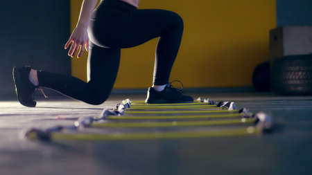 close-up, female legs in black leggings and sneakers. woman performs exercises with an athletes foot ladder, jumps , training on agility ladder on floor, in gym. Fitness class cardio workout .の写真素材