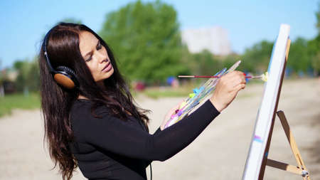 summer, near river, on beach. at sunrise, a beautiful woman artist holds a brush in her hand, draws a picture. she paints on canvas god Shiva. listening to music on headphones. High quality photoの写真素材