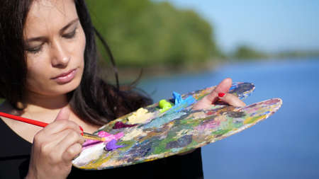 summer,outdoors, portrait of a beautiful forty years old brunette woman artist, close-up of a palette with paints, the woman artist mixes paints with a brush on the palette. High quality photoの写真素材