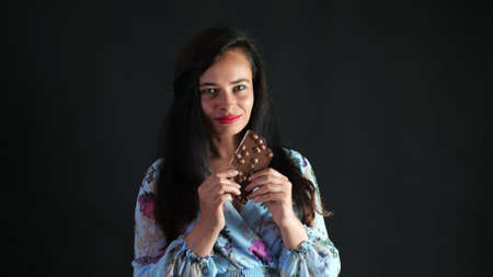portrait of a sexy brunette woman with red lips who eroticly and playfully eats black chocolate with hazelnuts and looking at camera in studio. dark gray background. High quality photoの写真素材