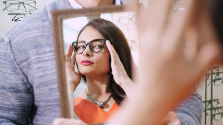 Young woman checking her new glasses in a mirror at optician retail store. Young Woman with...