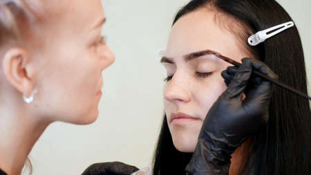 close up. young woman gets eyebrow correction procedure. kosmetolog- makeup artist applies paint...