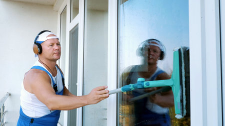 Caucasian attractive man, cleaning service worker, in blue overalls, listening to music on mobile...