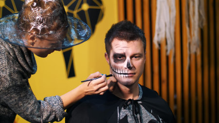 Halloween party, make-up artist draws a terrible makeup on the face of a man for a Halloween party. in the background, the scenery in the style of Halloween is seen. High quality photoの写真素材