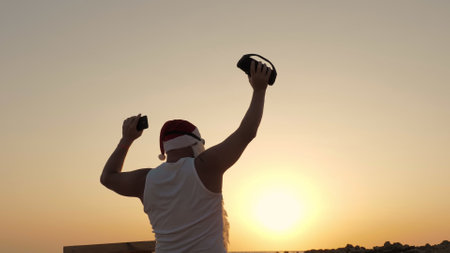 Santa Claus dances. Funny Santa Claus dancing with a wireless musical wireless speaker in hand, against backdrop of sunset or sunrise. back view. relax on beach by the sea. High quality photoの写真素材