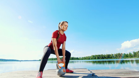 Beautiful, athletic young blond woman doing different exercises with weights, dumbbells, lunges, squats. Lake, river, blue sky and forest in the background, summer sunny day. High quality photoの写真素材