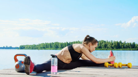 Beautiful, athletic young blond woman, coach, instructor, stretching, doing different exercises. Lake, river, blue sky and forest in the background, summer sunny day. High quality photoの写真素材