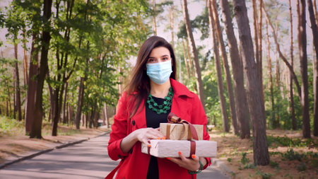 wrapped gifts. a woman in a protective mask holding several beautifully packed boxes in her hands. outdoors. delivery or donation, charity concept. gifts for the holiday. High quality photoの写真素材