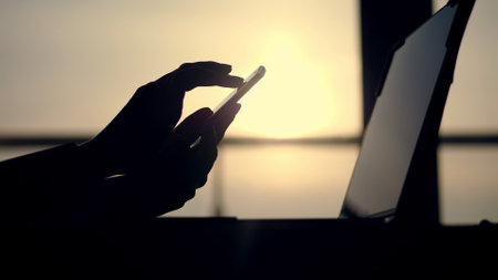 dark silhouette of female hands, typing something in mobile, next is laptop on table. against background of a large office window, at sunset, in the rays of light. close up. High quality photoの写真素材