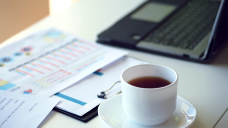 close up, on a white desktop there is a white cup of tea, next to a laptop and spread out business graphs. High quality photoの写真素材