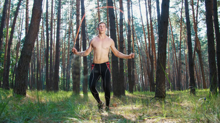 A young athletic man with a bare, naked torso, jumping rope, performs strength exercises with a rubber rope, a crossfit element. Slow motion. In pine forest, summer, in rays of sun. High quality photoの写真素材