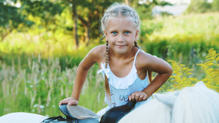 Blond girl of seven years, in a white dress, climbs, sits on a pony. Cheerful, happy family vacation. Outdoors, in the summer, near the forest. High quality photoの写真素材