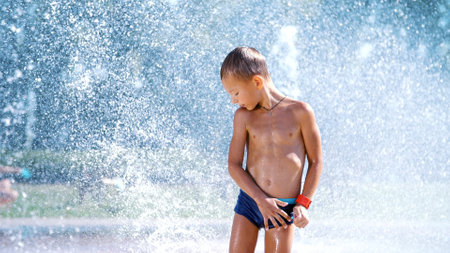 excited boy of seven years having fun between water jets, in fountain, run around, sprinkle, have fun, have fun, on a hot summer day. Summer in the city. Slow motion. High quality photoの写真素材
